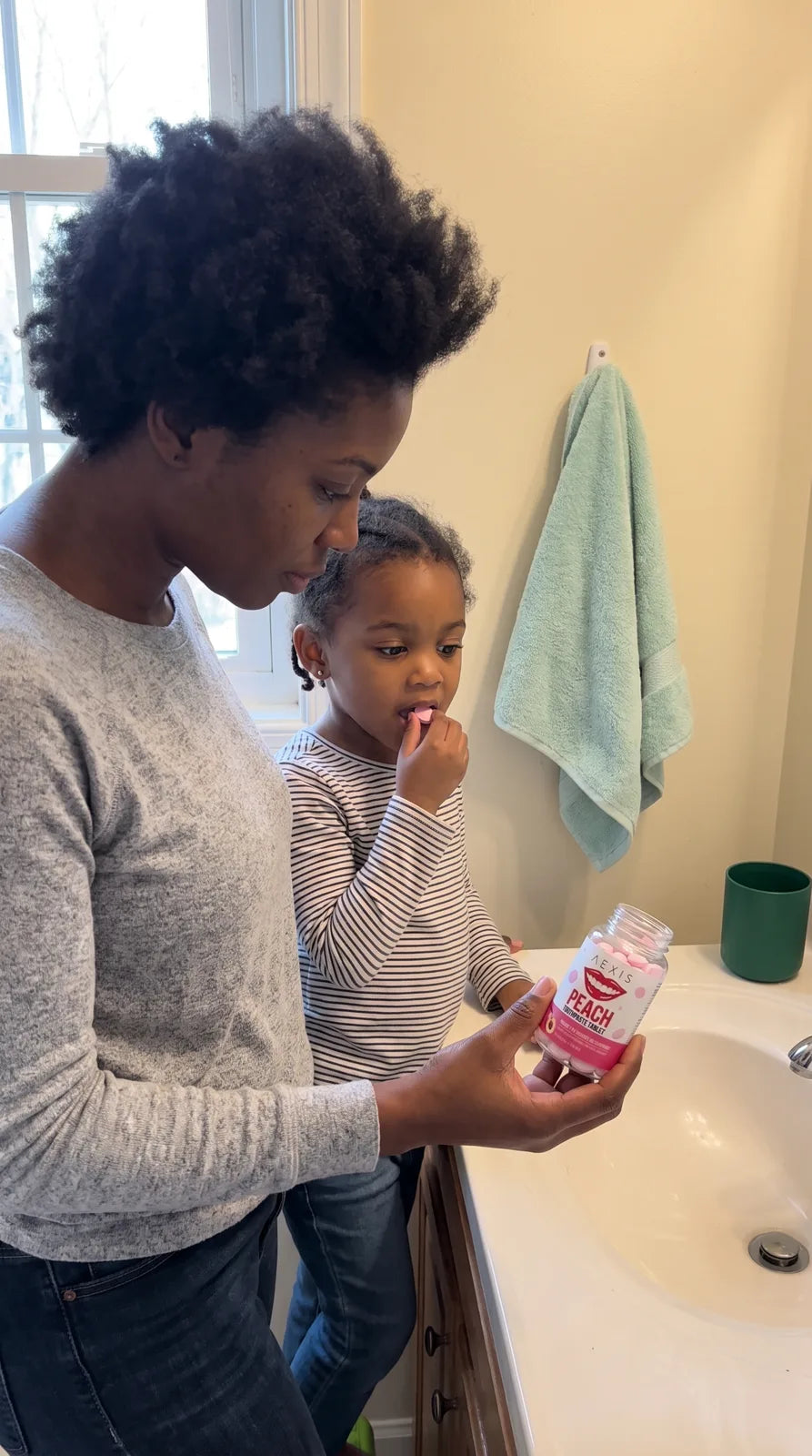 A girl chews a peach tablet at the bathroom sink beside her mom